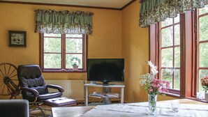 Quarto com televisão em móvel, uma poltrona de couro, mesa com um vaso de flores e janelas amplas com cortinas.