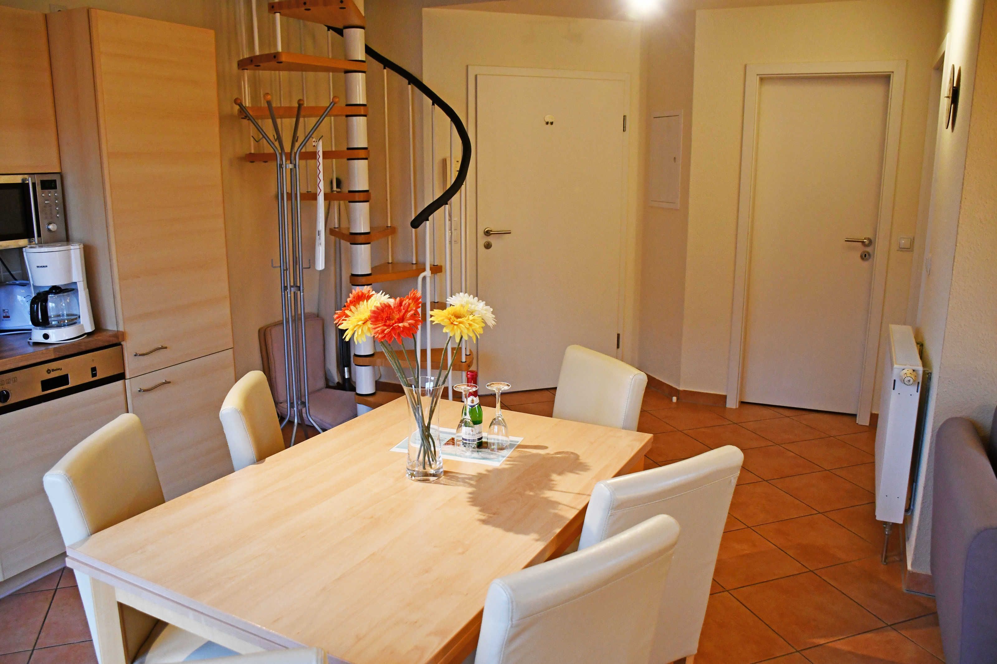 A dining area with a wooden table, white chairs, a vase with flowers, a spiral staircase, and a microwave.