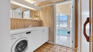 Een kleine keuken met een ingebouwde wasmachine, houten lambrisering, een wit aanrechtblad en uitzicht op een zwembad door een open deur.