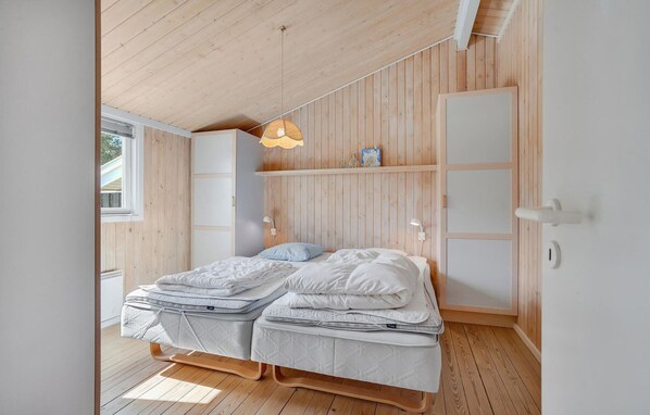 Un dormitorio con estructura de cama de madera, dos colchones blancos, una mesita de noche, una lámpara colgante y una ventana con persianas.