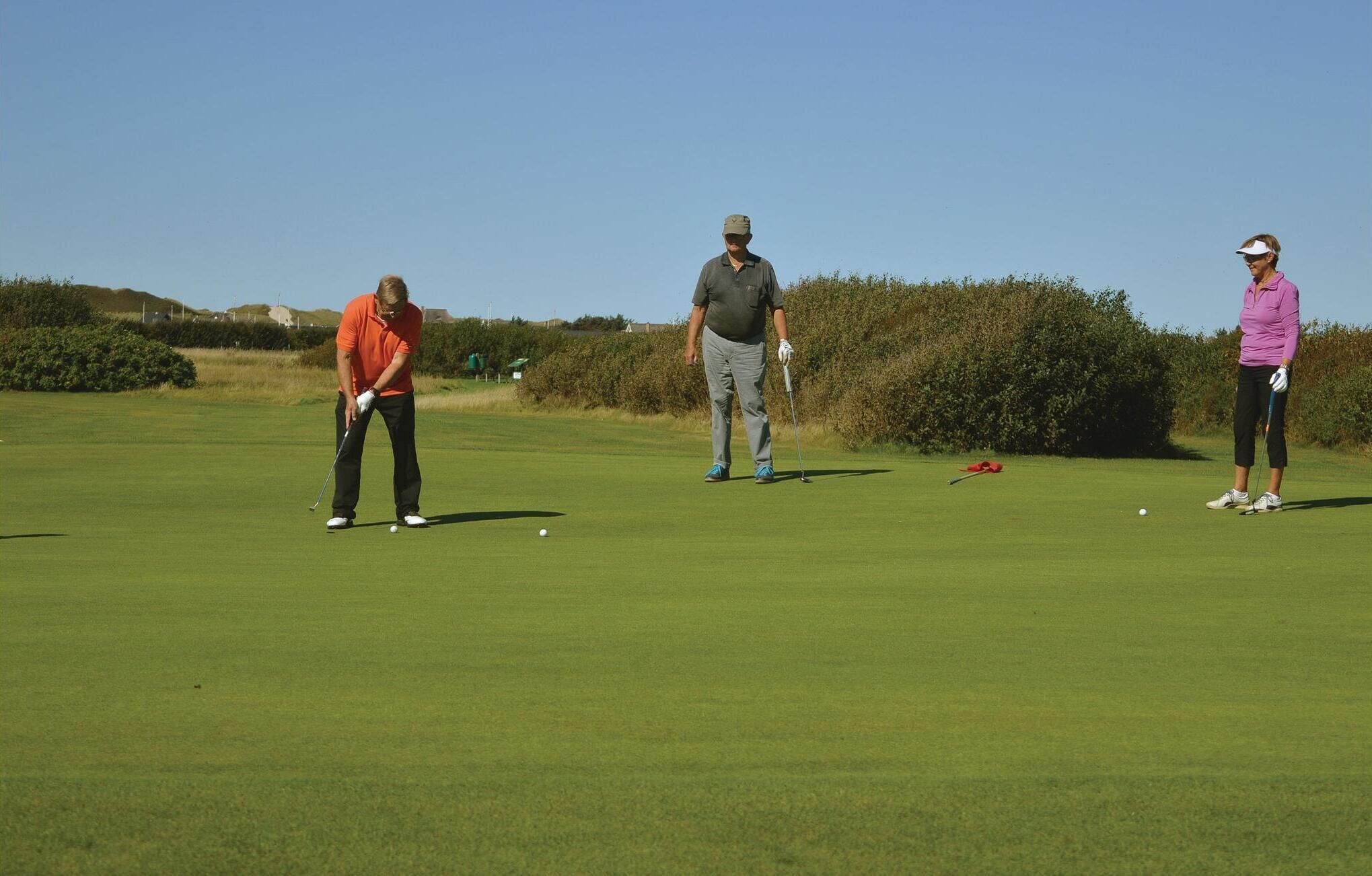 Un terrain de golf où trois personnes jouent, un ciel dégagé et une haie en arrière-plan.