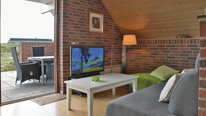Una habitación acogedora con pared de ladrillo, televisor, mesa blanca, sofá gris y un pequeño balcón con mobiliario de mimbre.