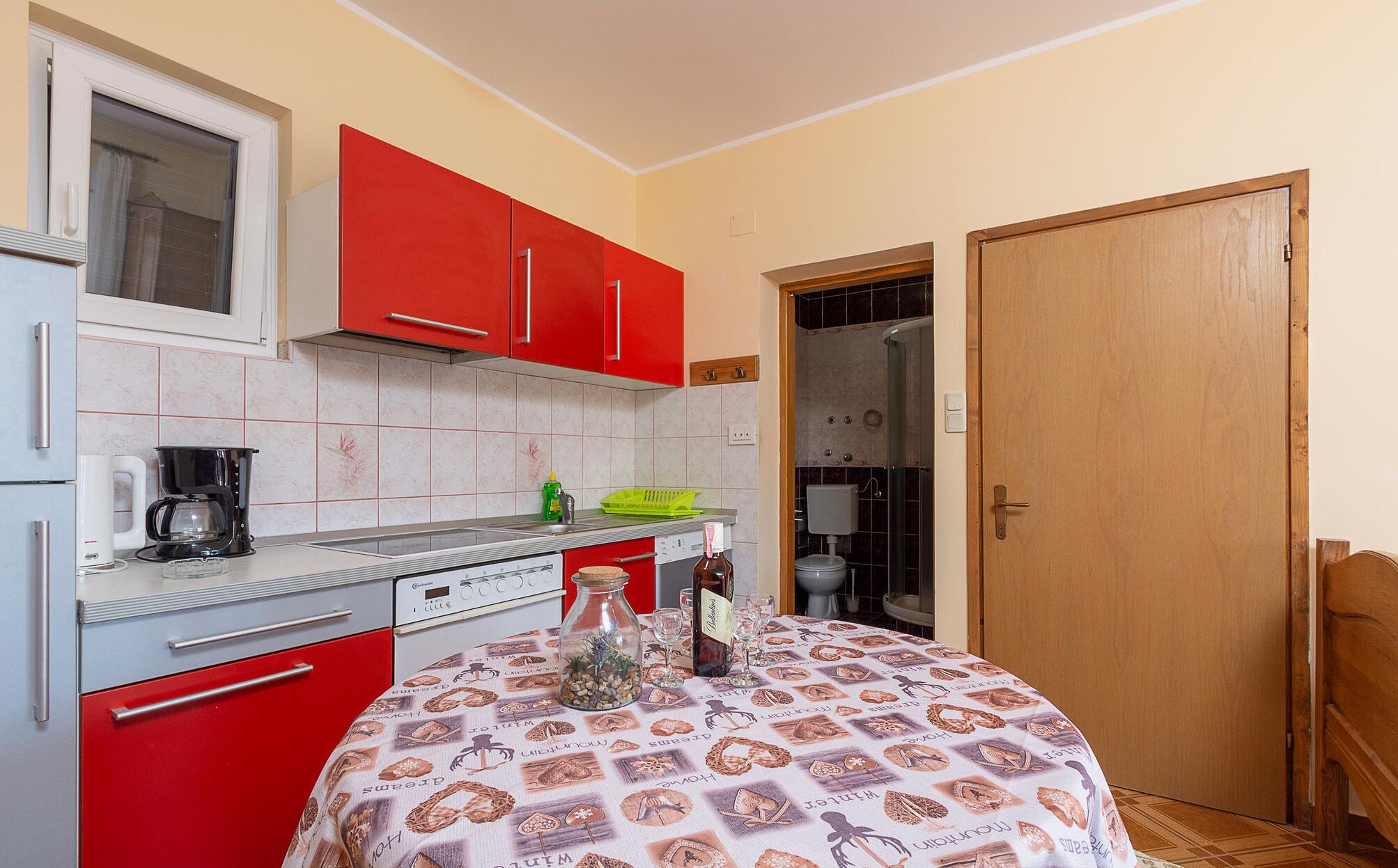 Une cuisine de taille réduite avec des placards rouges, une machine à café et une table recouverte d’une nappe à motifs.
