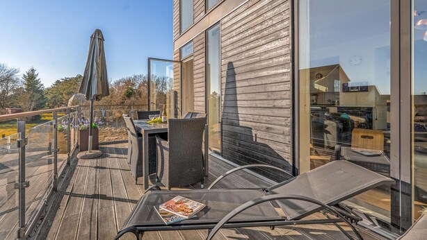 Een modern terras buiten met een glazen balustrade, rieten meubels en uitzicht op het omliggende landschap.