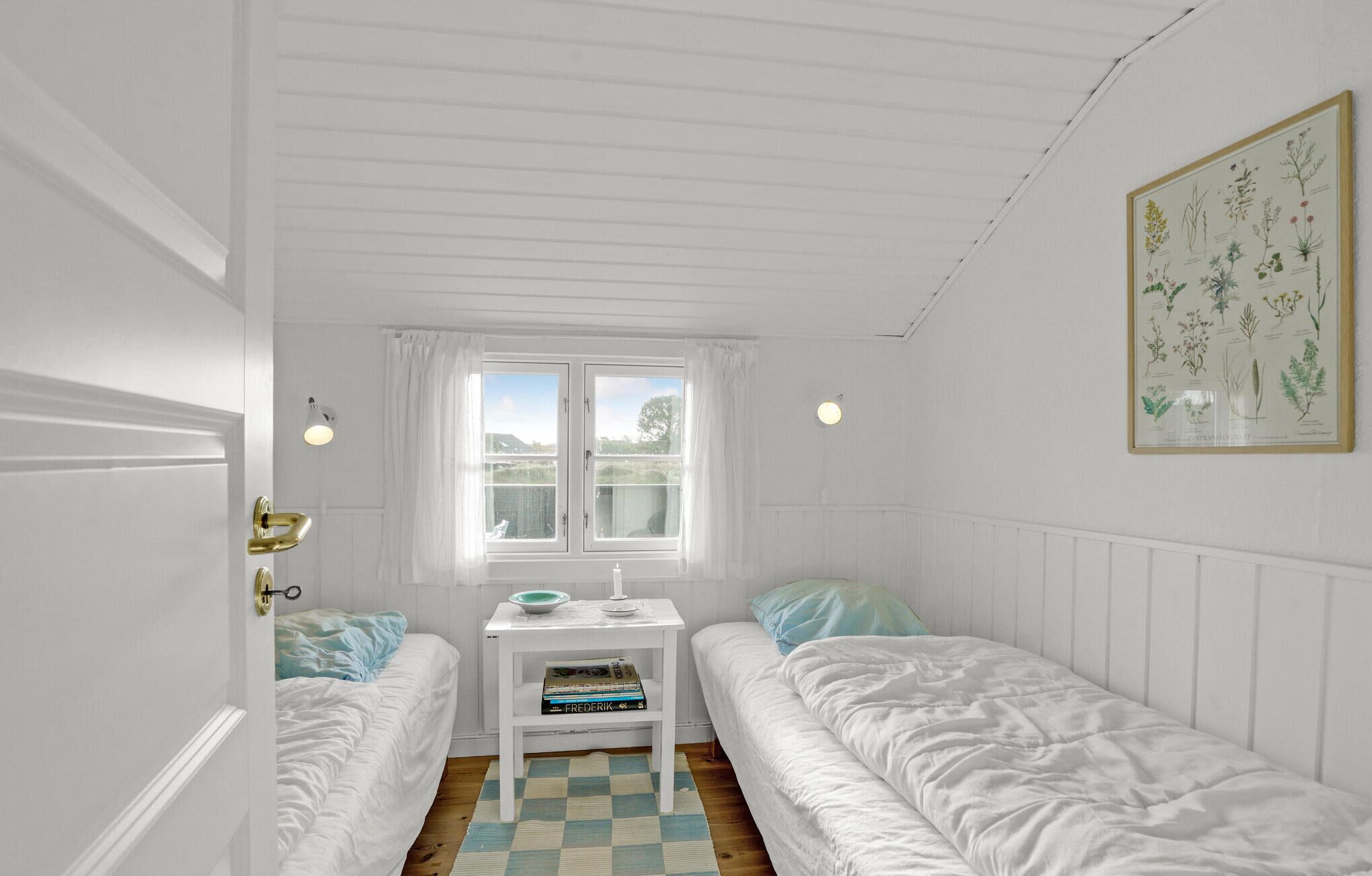 A small, white room with two single beds, a small table with a bowl and a book, a window with curtains, and a framed botanical illustration on the wall.