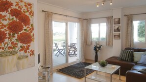 Un salon comprenant un canapé, une table basse, un téléviseur et un tableau représentant des fleurs. On y trouve également des rideaux, une fenêtre et une porte donnant sur un espace extérieur aménagé avec des chaises.