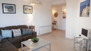 Un salon avec un canapé en cuir marron, une table basse en verre et un téléviseur posé sur un meuble. Les murs sont ornés de tableaux encadrés et un radiateur se trouve dans un coin.