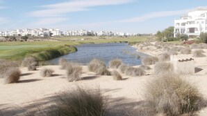 Une rivière aux rives sablonneuses et à la végétation clairsemée, bordée d’un terrain de golf et entourée de bâtiments résidentiels en arrière-plan.
