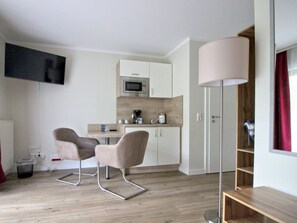 Ein modernes Zimmer mit Flachbildfernseher, einer Küchenzeile, zwei Stühlen, einer Stehlampe und einem Holzregal.