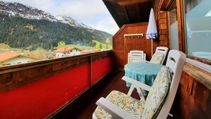 Un balcón con mesa y sillas, un muro de madera y vistas a montañas y casas.
