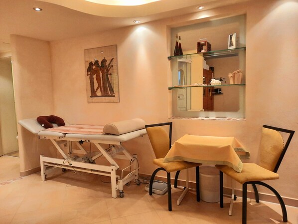 Un consultorio médico con una camilla, una pequeña mesa con sillas, un estante en la pared con objetos personales y una obra de arte enmarcada.
