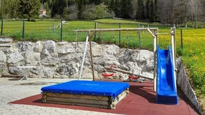 Un parque infantil con un tobogán y un tapete azul, rodeado por un muro de piedra y una zona vallada con montañas al fondo.