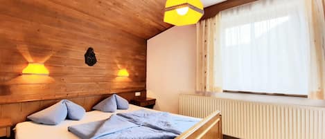 A wooden room with a bed, two wall-mounted lamps, a radiator, and a window with curtains.