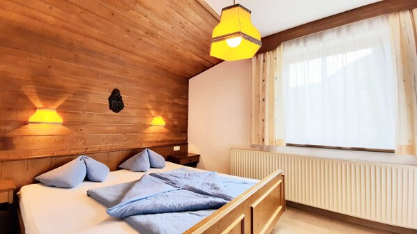 A wooden room with a bed, two wall-mounted lamps, a radiator, and a window with curtains.