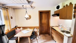 A compact kitchen with a dining table, chairs, a hanging light fixture, wooden cabinets, and a door leading to another room.