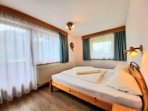 A bedroom with a wooden bed, white bedding, a radiator, and two windows with curtains.