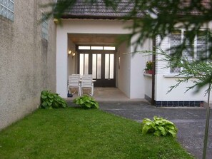 Ein Gebäudeeingang mit einer Tür, zwei weißen Stühlen und einem kleinen Garten mit Pflanzen.