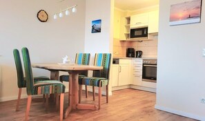 Uma área de refeições com mesa e cadeiras de madeira, uma cozinha com armários e eletrodomésticos brancos, e um relógio fixado na parede.