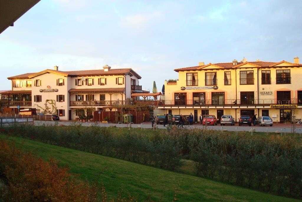 A row of buildings with multiple stories, some with balconies, and a few with signage indicating businesses. There are parked cars in front of the buildings.