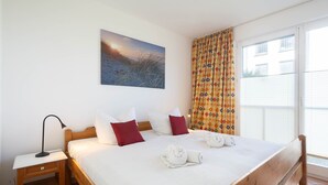 A bedroom with a wooden bed, white bedding, a nightstand, a lamp, a painting on the wall, and a window with curtains.
