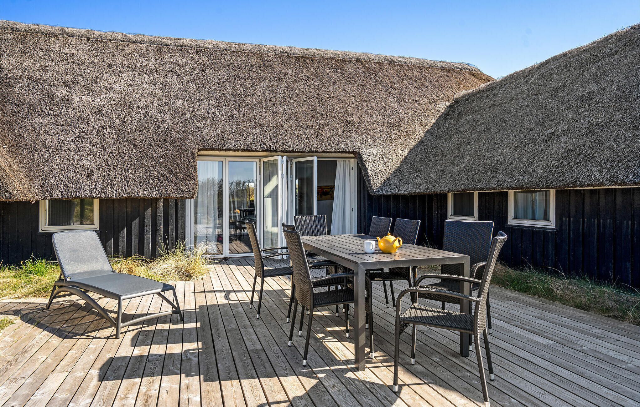 Een houten terras met een tafel en stoelen, een gebouw met rieten dak en een glazen deur die leidt naar de binnenruimte.