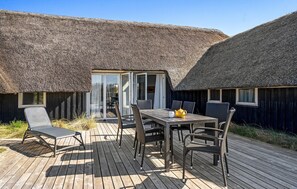 A wooden deck with a table and chairs, a thatched-roof building, and a glass door leading to an interior space. - Lovely home in Blåvand with sauna (Blåvand)