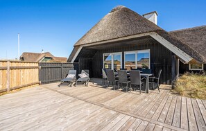 A wooden deck with a thatched-roof building, featuring a dining area with chairs and a table. - Lovely home in Blåvand with sauna (Blåvand)