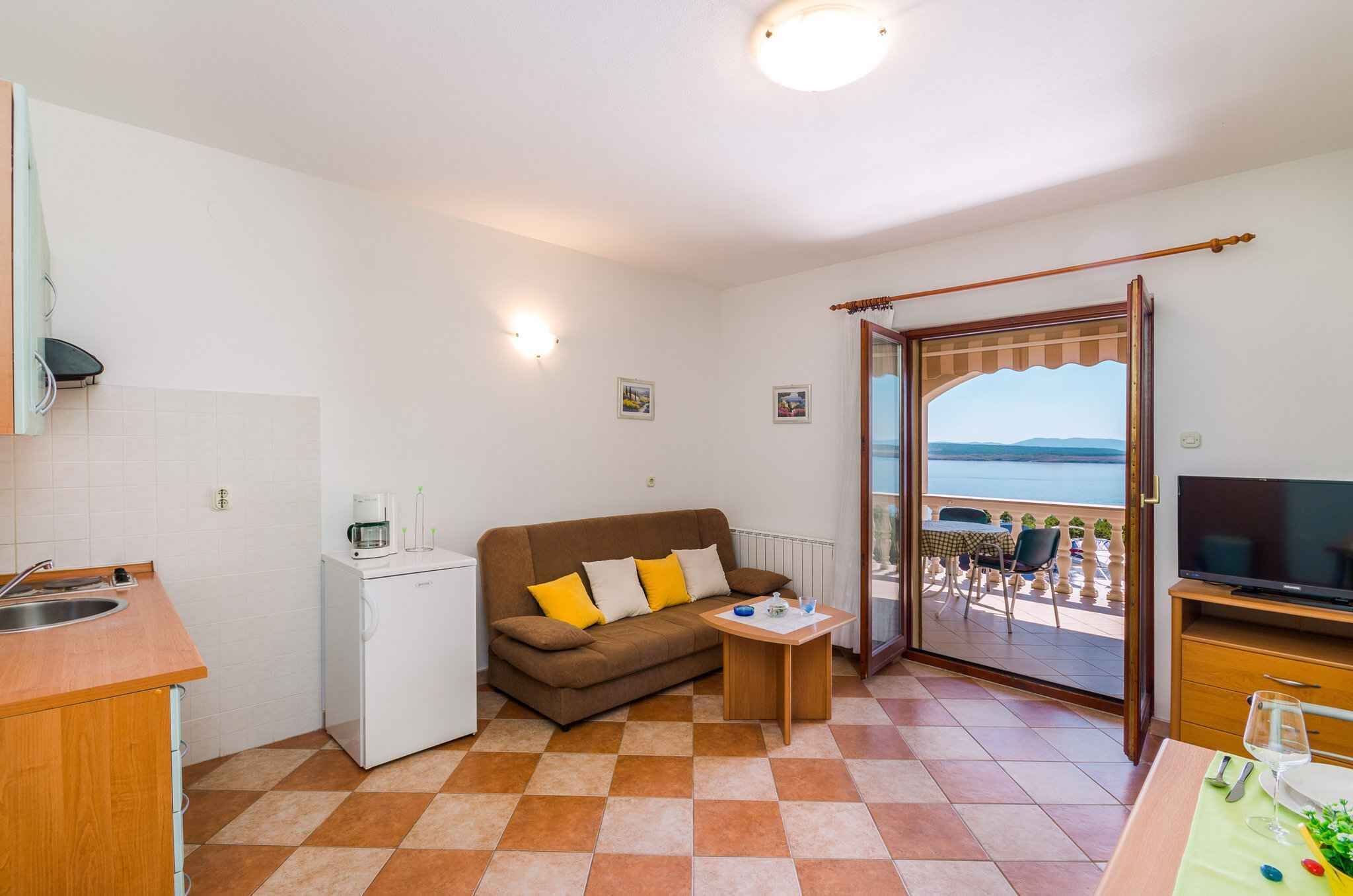 Ein kompakter Wohnraum mit Küchenzeile, einer Couch mit Kissen, einem kleinen Tisch und einem Balkon mit Blick.