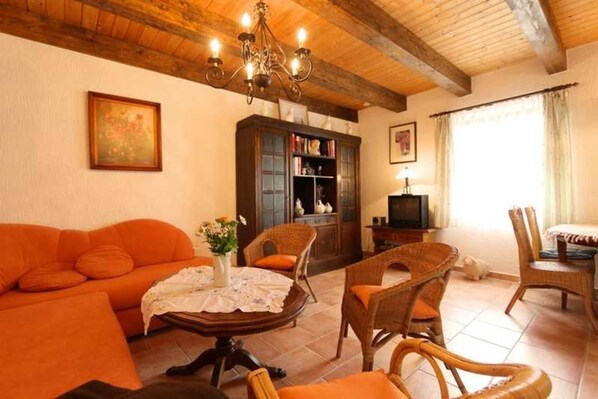 Een gezellige woonkamer met een houten plafond, een bank, rieten stoelen, een tafel met een vaas en een boekenkast.