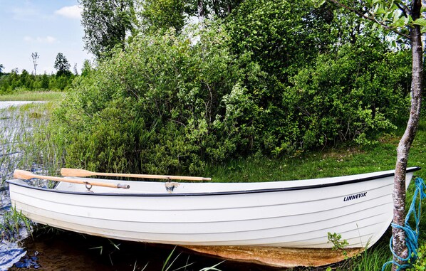 Un petit bateau blanc, avec une pagaie en bois posée dessus, est attaché à un arbre par une corde bleue. Le bateau est marqué du nom « LINNEVIK » et est amarré près d’une zone herbeuse, avec en arrière-plan un feuillage vert et dense.