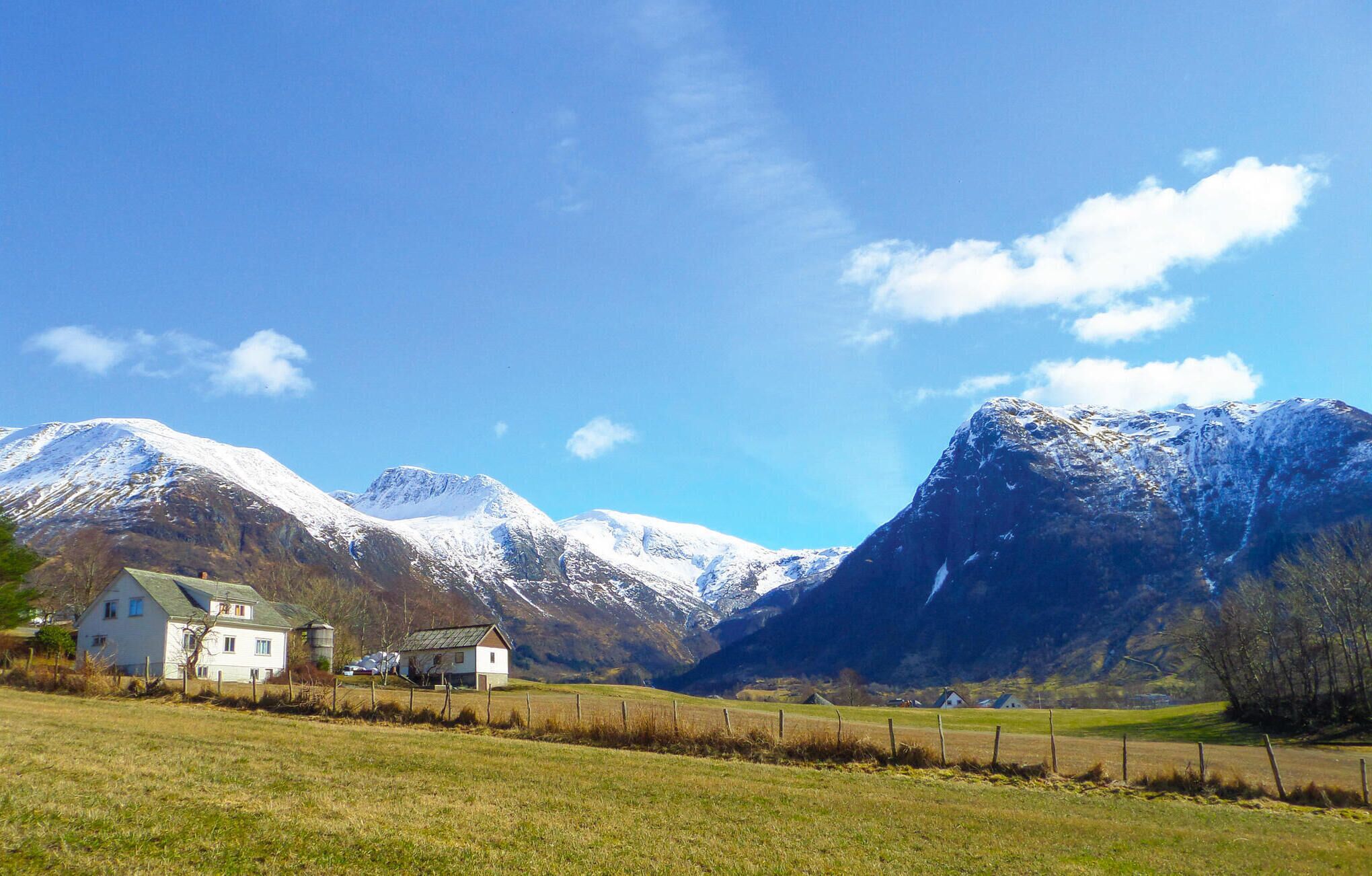 A rural landscape with a house and a barn, surrounded by snow-capped mountains under a clear sky.