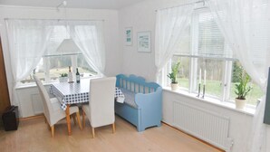 Ein Zimmer mit Esstisch, Stühlen, einer blauen Bank und großen Fenstern mit weißen Vorhängen.