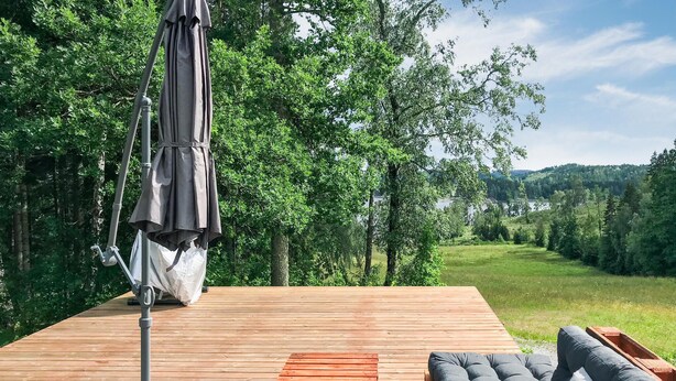 Eine Holzterrasse mit grauem Sonnenschirm, einer gepolsterten Sitzecke und Blick auf eine bewaldete Landschaft.