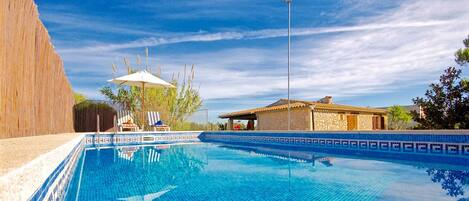 A swimming pool with a tiled edge, a covered seating area with chairs, and a stone building in the background.