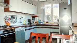 Une cuisine équipée d’une cuisinière à gaz, d’un lave-vaisselle et d’un réfrigérateur. Une fenêtre avec des rideaux offre une vue sur les arbres à l’extérieur.