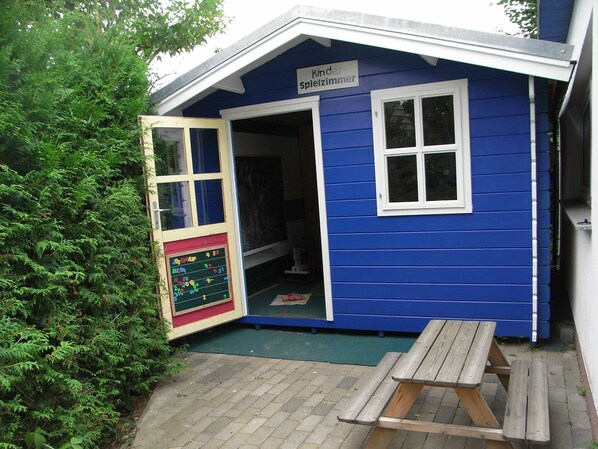 Une salle de jeux bleue avec une pancarte indiquant « Kinder-Spielzimmer », un tableau noir et un banc en bois à l’extérieur.