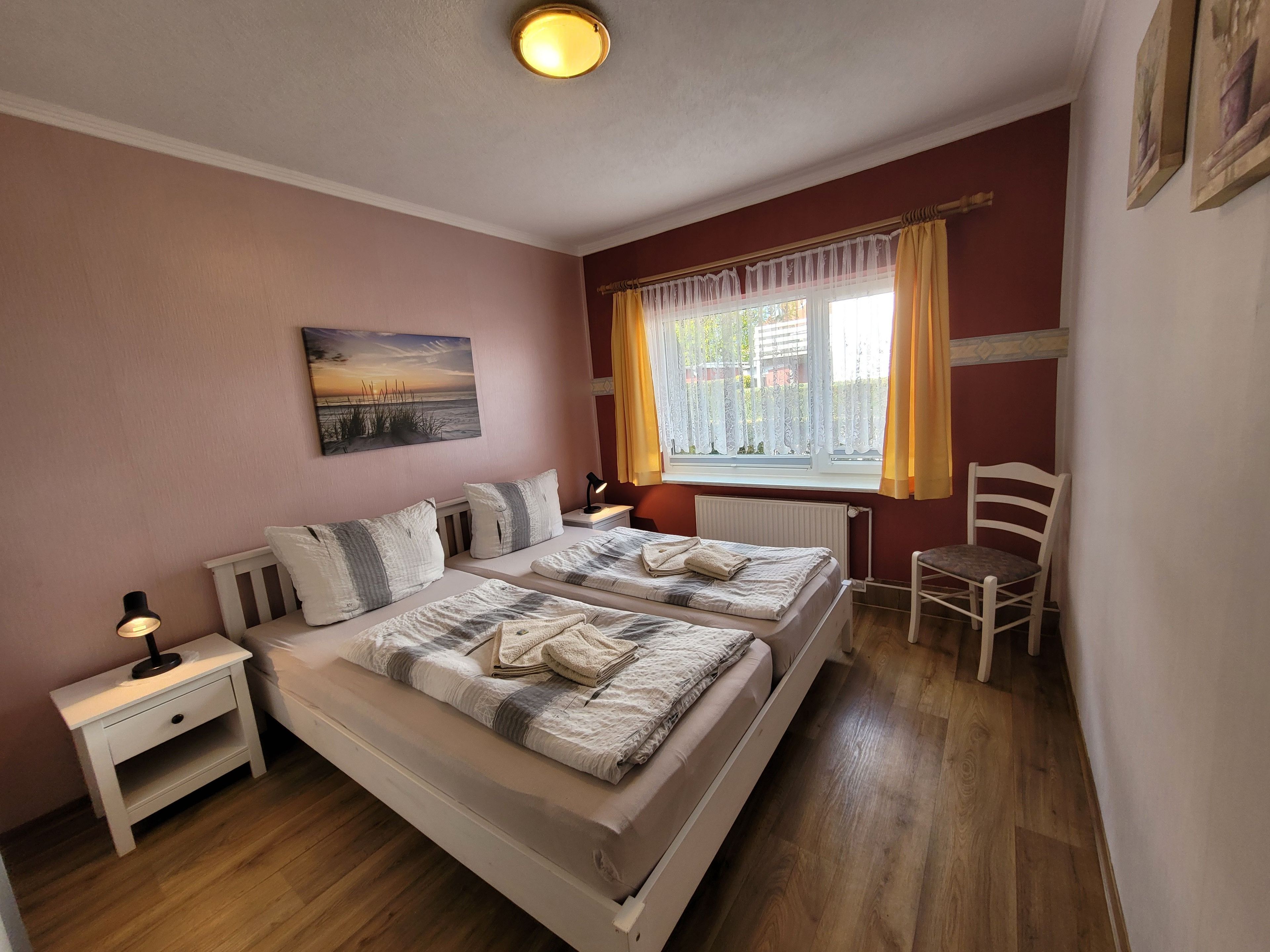Une chambre avec deux lits, une table de chevet, une chaise, une fenêtre avec des rideaux et un tableau au mur.