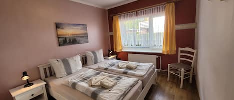 Un dormitorio con dos camas, una mesita de noche, una silla, una ventana con cortinas y un cuadro en la pared.