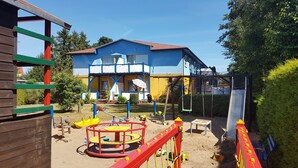 Un'area giochi con altalene e scivolo, una struttura in legno per bambini e una casa sullo sfondo.