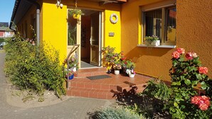 Un edificio giallo con tetto rosso, una porta e una finestra. Intorno all'ingresso, piante e fiori.