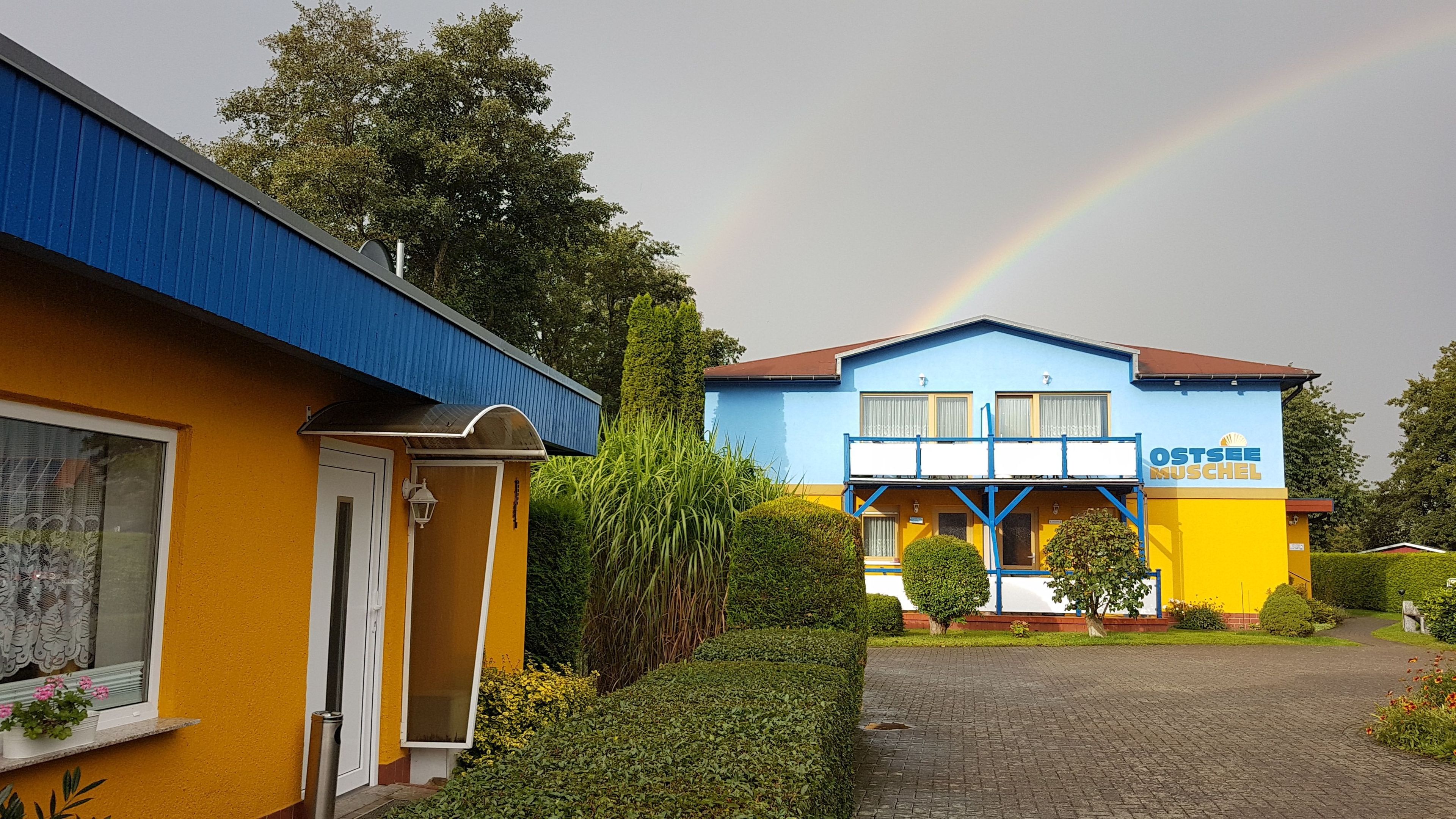 Un hotel con fachada azul y amarilla, un arcoíris visible y un camino de entrada pavimentado.