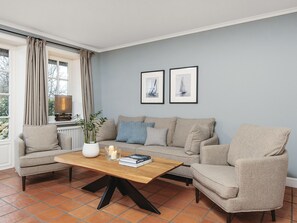 Een woonkamer met een beige bank, een houten salontafel en een stoel. Twee ingelijste foto’s hangen aan de muur, er staat een lamp op een tafel en een plant in een witte vaas.