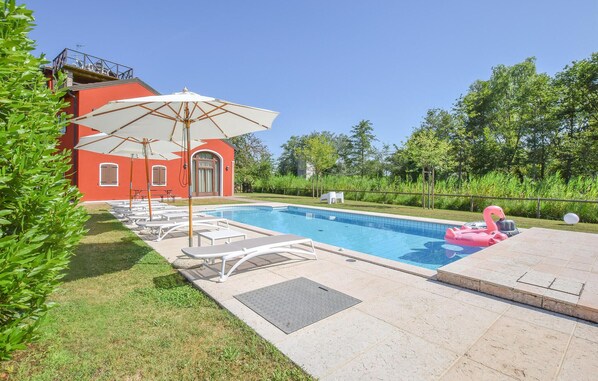 Ein Swimmingpool mit Liegestühlen und einem Sonnenschirm, umgeben von Grünpflanzen und einem roten Gebäude.