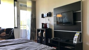 Habitación de hotel con una cama, televisor montado en la pared, una mesita con un altavoz y una estantería con revistas.