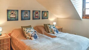 A bedroom with a sloped ceiling, a bed with a white quilt, two nightstands, and four wall-mounted paintings of dogs on chairs.
