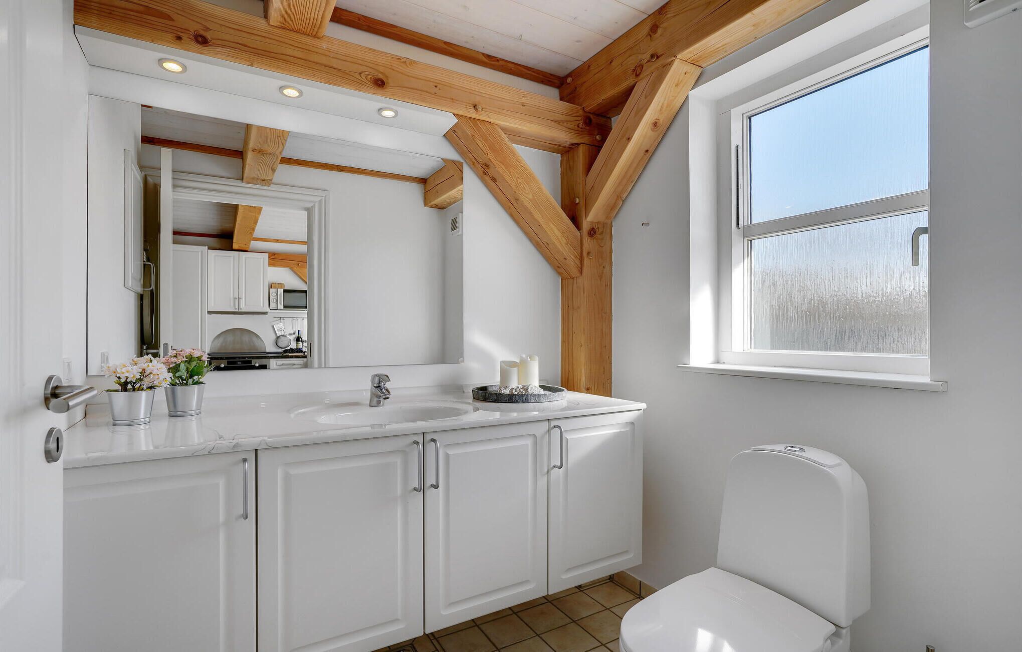 A bathroom with a white sink, mirror, and toilet, wooden beams on the ceiling, and a window.