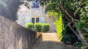 A stone building with a window and shutters, surrounded by greenery and a gravel path.