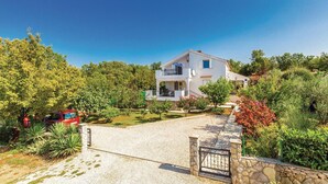 Une maison à deux étages dotée d’un balcon, entourée d’une végétation luxuriante et accessible par une allée de gravier.