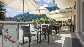 Ein Straßencafé mit Korbstühlen und großen Sonnenschirmen mit Blick auf eine Berglandschaft.
