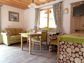 A cozy living room with a wooden ceiling, a green sofa, a dining table with chairs, and a television on the wall.
